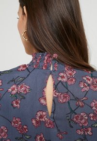 Volná modrá halenka s květinovou výšivkou ve růžové a zelené barvě. Nařasený vysoký límec a otvor ve tvaru klíče na zadní straně pro přidanou zajímavost.