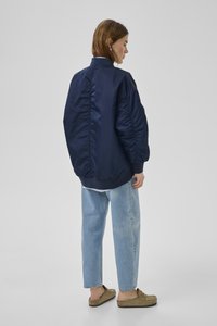 Frau im Profil trägt eine übergroße marineblaue Jacke, hellblaue Jeans und braune Slipper vor schlichter grauer Hintergrund.