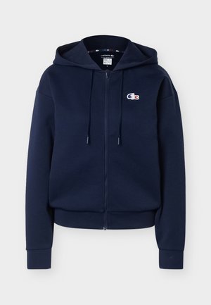 Marineblå hettegenser med glidelås, snorer, ribbestrikkede mansjetter og hem, samt en liten brodert Lacoste-krokodillelogo på venstre bryst.