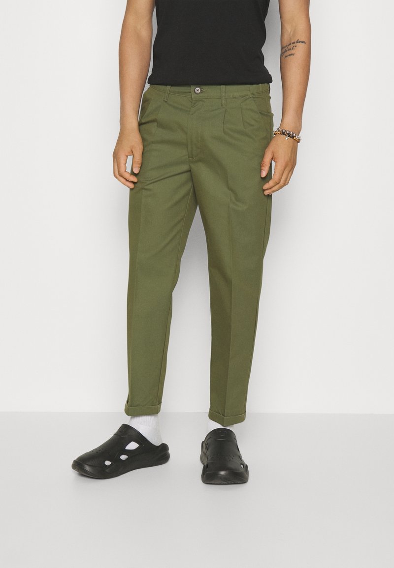 Pantalon vert olive avec une coupe droite, doté de poches avant et d'une fermeture par bouton, associé à des sabot noirs et des chaussettes blanches.