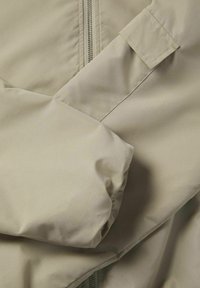 Beige pufferjack detail met een ritsafsluiting, wijde mouwen en een gladde, matte textuur met zichtbare stiksels.
