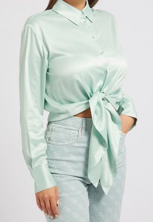 Femme portant un chemisier en satin vert clair boutonné et noué devant, associé à un jean bleu clair à motifs.