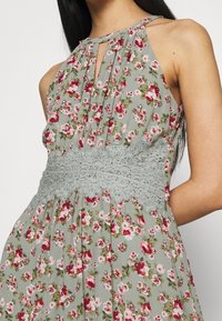 Robe fleurie en tissu vert clair avec des roses rouges et roses, dotée d'un col en trou de serrure et d'un détail en dentelle grise à la taille.
