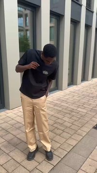 Schwarzes T-Shirt mit kurzen Ärmeln und Grafik auf der Brust, beiges plissiertes Hosen, und schwarze Schuhe, stehend auf einer steinernen Oberfläche.