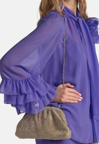 Pochette dorée à sequins avec chaîne, design froncé, tenue contre un chemisier en chiffon violet à manches volantées.