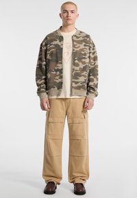 Camo bomberjack met een khaki basis, gecombineerd met beige cargobroeken met meerdere zakken, en bruine veterschoenen.