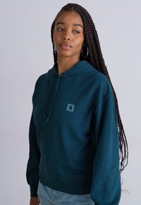 Hoodie bleu canard avec une poche kangourou et un capuchon à cordon réglable. Présente un patch logo carré dans le coin supérieur gauche. Texture lisse.
