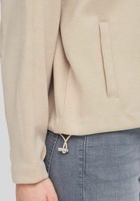 Felpa beige con una texture liscia, dotata di una tasca laterale e di un orlo regolabile con cordoncino e fermaglio metallico. Jeans grigi in vista.