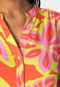 Kurzärmlige Bluse in lebendigem Pink, Orange und Grün mit Blumenmuster, ausgestattet mit einem kenten Kragen und einer Knopfleiste. Glatte Stoffstruktur.