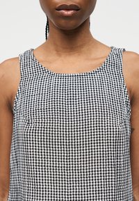 Κοντινό πλάνο ενός προσώπου που φοράει ασπρόμαυρη καρό αμάνικη μπλούζα με πλεξούδες στα μαλλιά και ουδέτερο χρώμα στα χείλη, σε απλό φόντο.