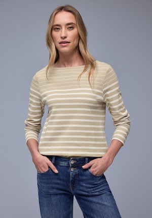 Donna con capelli biondi che indossa una maglia a maniche lunghe beige con strisce bianche e jeans blu, in piedi con le mani nelle tasche davanti a uno sfondo grigio.