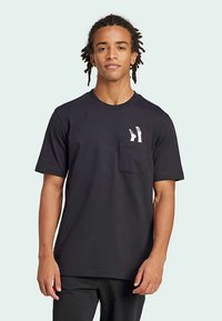 Νεαρός άντρας με πλεξούδες, φορώντας μαύρη μπλούζα με μικρή λευκή γραφική παράσταση δύο ποδιών πάνω από την τσέπη του στήθους.