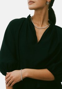 Sølv halskæde og armbånd med cirkulære elementer; modellen bærer en sort, tekstureret bluse med puffede ærmer og v-udskæring.