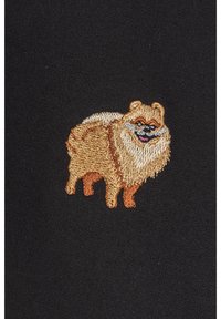 Broderad design av en fluffig pomeranian i ljusbrunt och krämvitt på en svart tygbakgrund. Har detaljerade ansiktsuttryck.