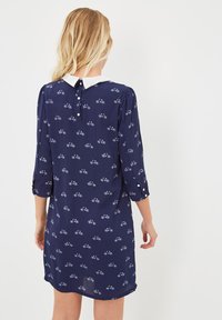 Mujer rubia vestida con un vestido azul marino con estampado de bicicletas blancas, cuello blanco y mangas de 3/4, de pie de espaldas sobre un fondo blanco.