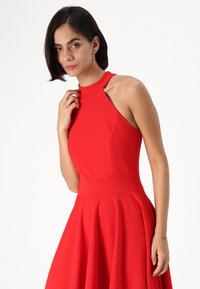 Rochie roșie fără mâneci, cu guler înalt, corset ajustat, fustă evazată și textură netedă. Include un design cu decupaje la umeri.