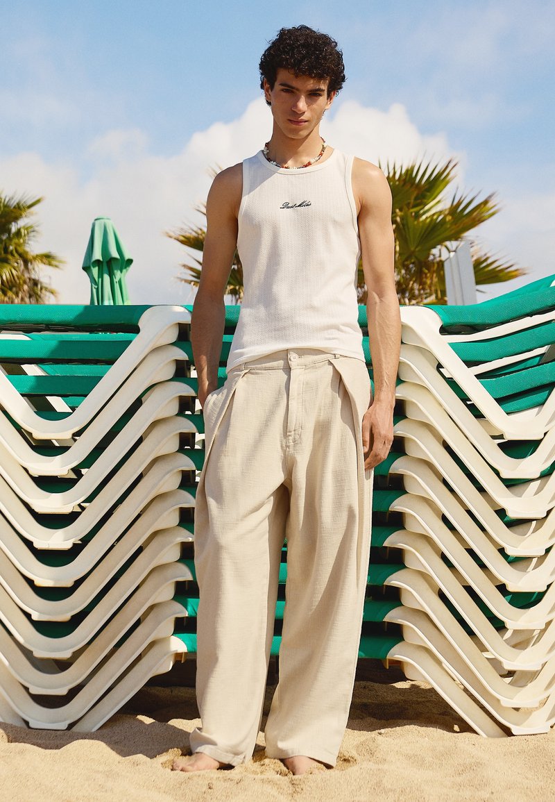 Jeune homme debout pieds nus sur le sable, portant un débardeur blanc et un pantalon beige ample devant des chaises de plage vertes et blanches empilées.