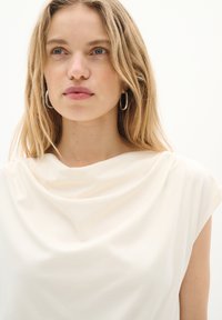 Haut à manches courtes de couleur crème avec un décolleté drapé ; tissu lisse et léger avec une texture douce et des détails de design minimaux.