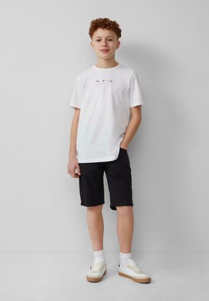 Biała bawełniana koszulka z grafiką, w połączeniu z czarnymi szortami. W zestawie białe sneakersy z beżową gumową podeszwą i skarpetami do kostek.