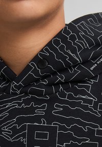 Sudadera negra con capucha que presenta un patrón abstracto blanco, con una tela texturizada y un detalle de botón a presión negro cerca del cuello.