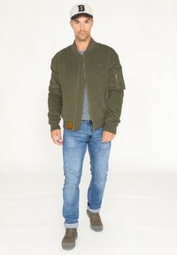 Veste bomber vert olive avec des poignets côtelés, des poches zippées et un tissu texturé, portée sur une chemise grise et un jean bleu, associée à des bottes beiges.