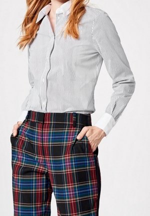 Mujer que lleva una camisa de botones blanca y gris a rayas metida en pantalones de cuadros de talle alto con las manos en los bolsillos.