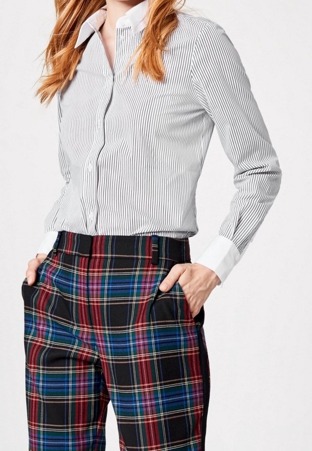 Femme portant une chemise blanche et grise rayée à boutons rentrée dans un pantalon à carreaux taille haute, les mains dans les poches.