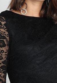 Svart spetsklänning med ett strukturerat blommönster, genomskinliga spetsärmar och en slät kropp. Inkluderar båtutskärning och rundade detaljer.