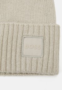 Šviesiai pilkas megztas kepurė su raukšlėta tekstūra ir stačiakampiu siuvinėtu ženkliuku, kuriame subtiliu tonu užrašyta „BOSS“. 