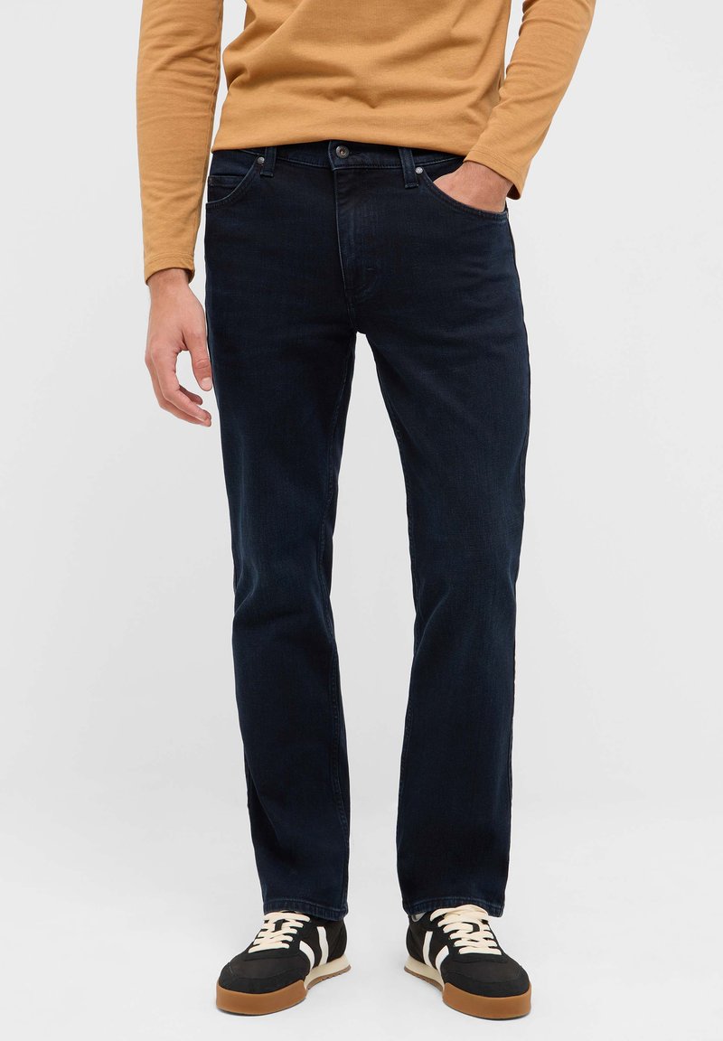 Dunkelblaue Slim-Fit-Jeans aus Denim, mit Vordertaschen und einer einzelnen Gesäßtasche, kombiniert mit schwarzen und cremefarbenen Turnschuhen.