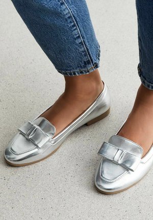 Srebrne metaliczne loafersy z akcentem w postaci kokardy, płaskim obcasem i gładkim wykończeniem, zestawione z niebieskimi dżinsami podwiniętymi na mankietach.