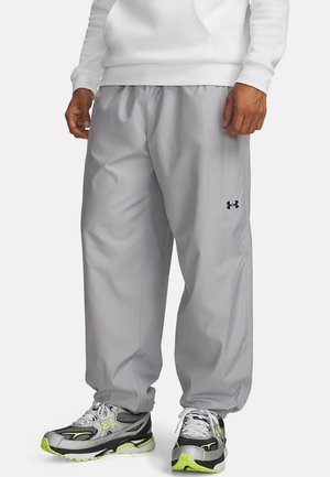 Man draagt lichtgrijze Under Armour trainingsbroek, witte hoodie en grijze, zwarte en neon groene sportschoenen, staand op een witte achtergrond.