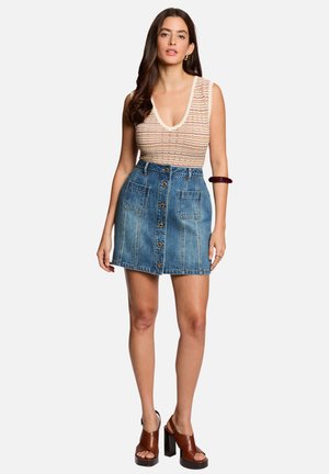 Femme aux longs cheveux foncés portant un haut sans manches à motifs, une jupe en jean boutonnée devant, des sandales marron à talons blocs et un bracelet sombre.