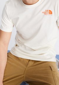 Krémové bavlněné tričko s krátkým rukávem. Na levém hrudi má malé oranžové logo. Nosené se světle hnědými kalhotami.