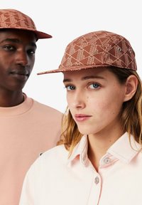 Cappello marrone a fantasia con visiera piatta. Il design presenta forme geometriche. Sullo sfondo si può vedere una camicia rosa chiaro con colletto dettagliato.
