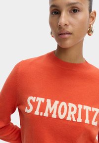 Pomarańczowy sweterek dziany z okrągłym dekoltem, z białym napisem "ST. MORITZ" wyraźnie wyeksponowanym na klatce piersiowej. Gładka tekstura.