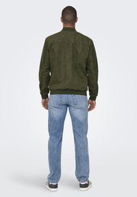 Giacca bomber in suede verde oliva con polsini e colletto a coste, abbinata a jeans in denim azzurro. Il retro presenta un design pulito e semplice.