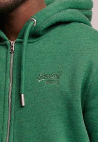 Felpa verde con zip realizzata in tessuto morbido, dotata di zip in metallo, coulisse e logo ricamato "SuperDry" sul petto.