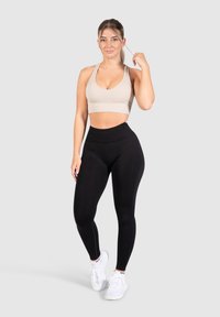 Beige sportbeha met geribbelde textuur, gecombineerd met hoge zwarte leggings. Model draagt witte sportschoenen. Eenvoudig, aansluitend ontwerp.