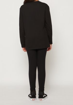 Person trägt ein schwarzes Langarmshirt, schwarze Leggings und schwarz-weiße High-Top-Sneaker und steht mit dem Rücken zur Kamera.