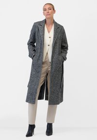 Grå texturerad överrock med en skalkrage, två framfickor och lång längd, i kombination med en krämfärgad topp och beige byxor. Svarta klackstövlar.