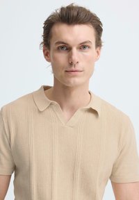 Jonge man met een lichte huid en blauwe ogen, gekleed in een beige, gestructureerd gebreide polo, recht vooruit kijkend tegen een eenvoudige witte achtergrond.