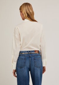 Chemise blanche à manches longues avec une coupe décontractée, ornée de broderies sur les manches, associée à un jean taille haute en bleu marine et une ceinture en corde.