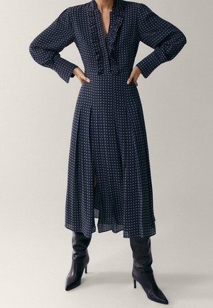 Femme portant une robe midi à manches longues sombre à motifs avec des volants et des bottes noires à talons hautes jusqu'aux genoux, debout les mains sur les hanches.