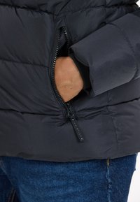 Svart puffjacka med ribbade manschetter, med en dragkedjeficka på sidan och en liten märkesetikett nära dragkedjans stängning.