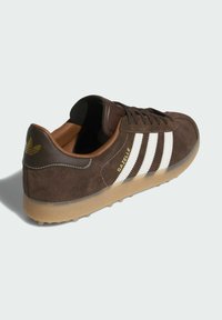 Sneakers en suede marron avec trois rayures blanches, talon en cuir marron, texte doré "Gazelle" et semelle en caoutchouc translucide.
