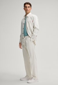 Witte denimjas met borstzak over een teal shirt, gecombineerd met crèmekleurige broek en witte sneakers. Knopdetails op de jas.