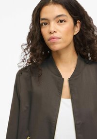 Brun bomberjakke med ribbestrikket krage, glidelåsfeste foran og sidelommer, laget av glatt stoff. Brukes over en hvit topp.