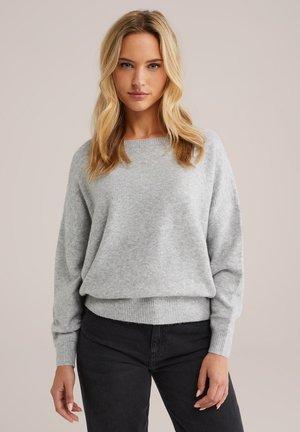 Hellgrauer Oversized-Pullover mit rundem Halsausschnitt und geripptem Saum. Weicher Stoff und überschnittene Schultern, kombiniert mit dunklen Jeans.