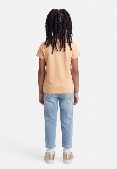 Παιδί με πλεξούδες, που φορά ροδακινί μπλούζα, ανοιχτό μπλε τζιν, λευκές κάλτσες και χρυσά αθλητικά παπούτσια, στέκεται γυρισμένο προς τα πίσω σε λευκό φόντο.